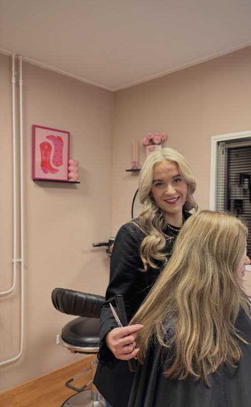 Luca van de Kuilen in haar salon, met kam en schaar naast een klant met lang blond haar